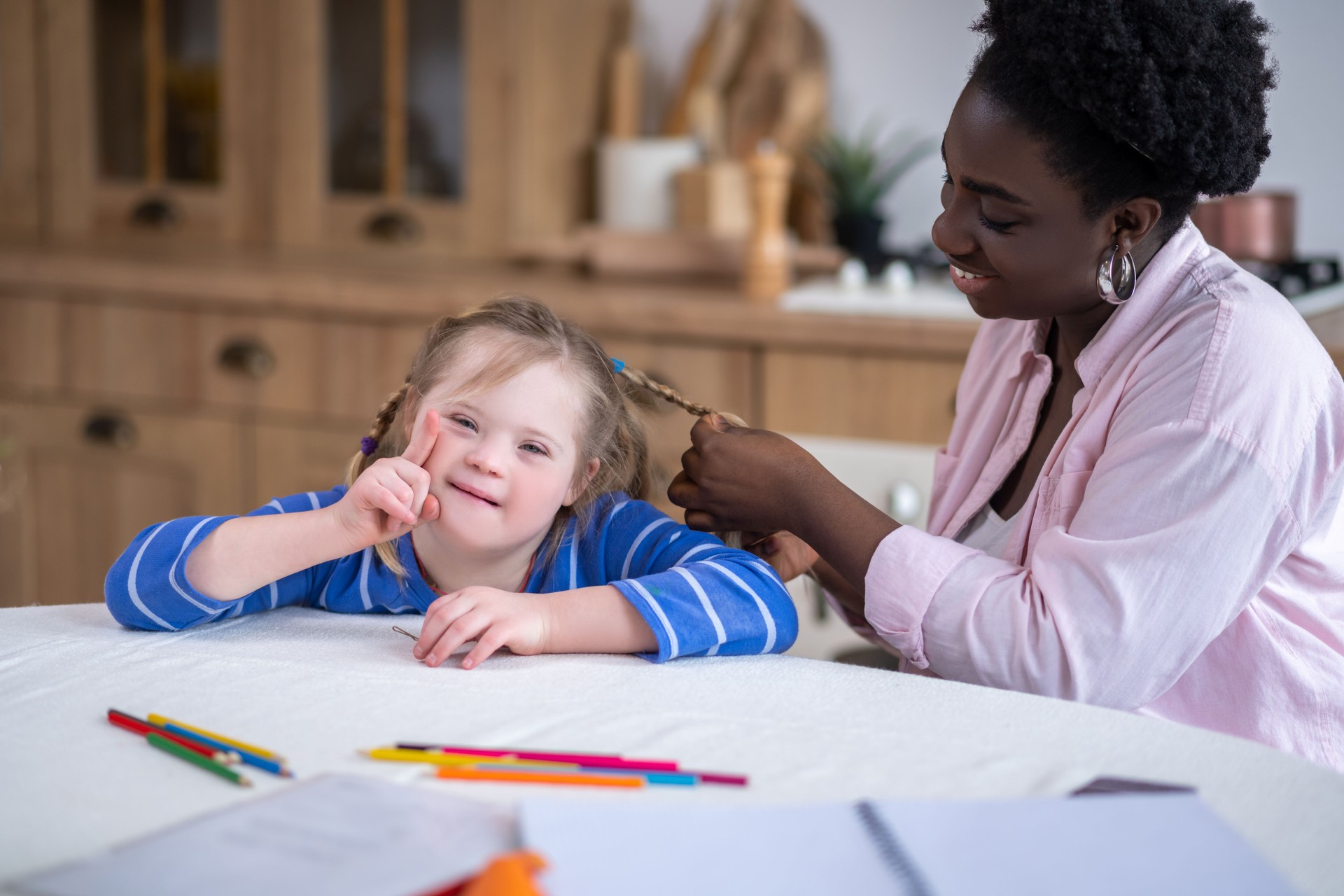 A smiling dark-skinned woman doing hair to the cute girl with down syndrome