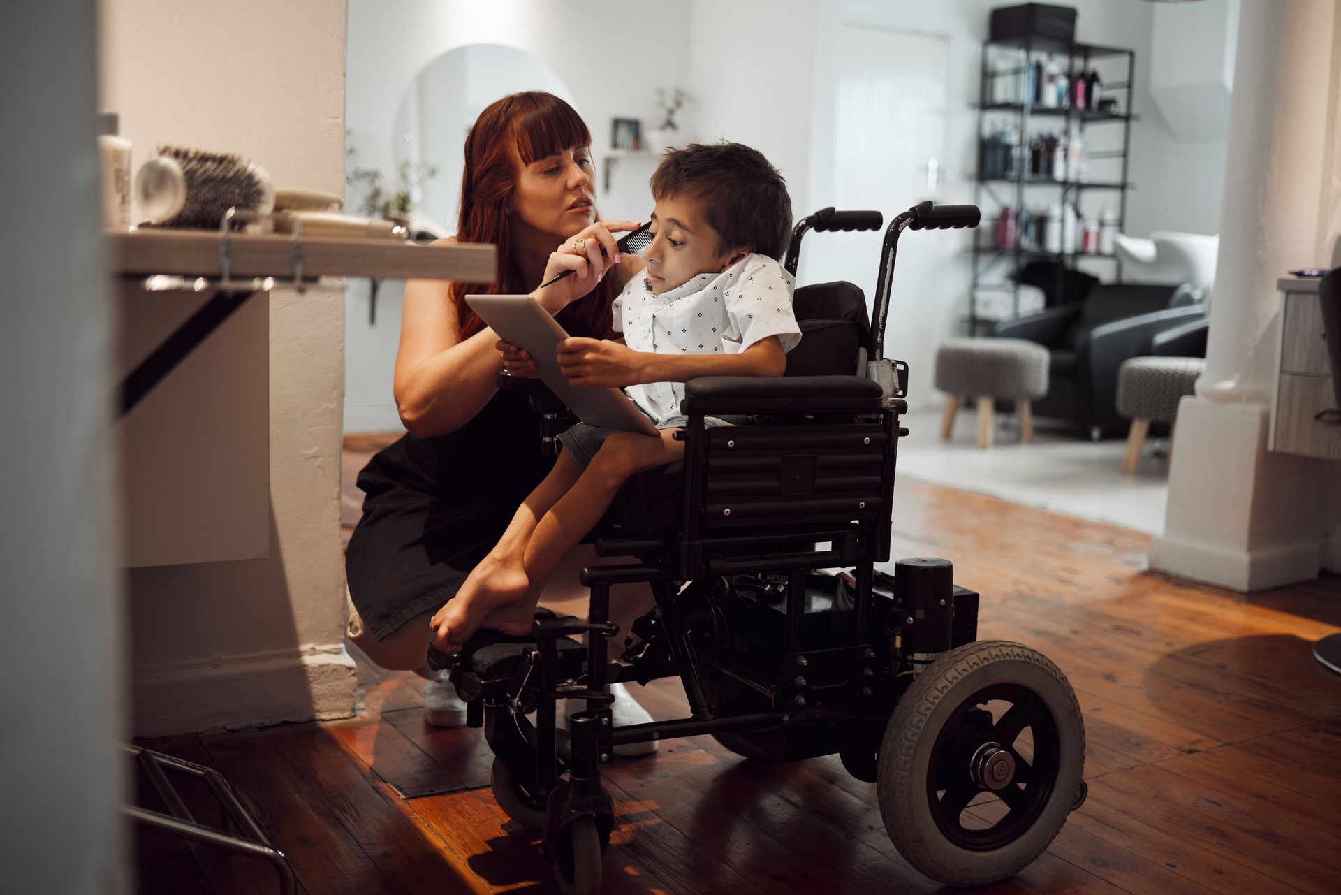 Hair, beauty and inclusive barber grooming child in a wheelchair, relax while digital tablet and comfort. Care, disability and child development with handicapped boy enjoying pamer treatment haircut